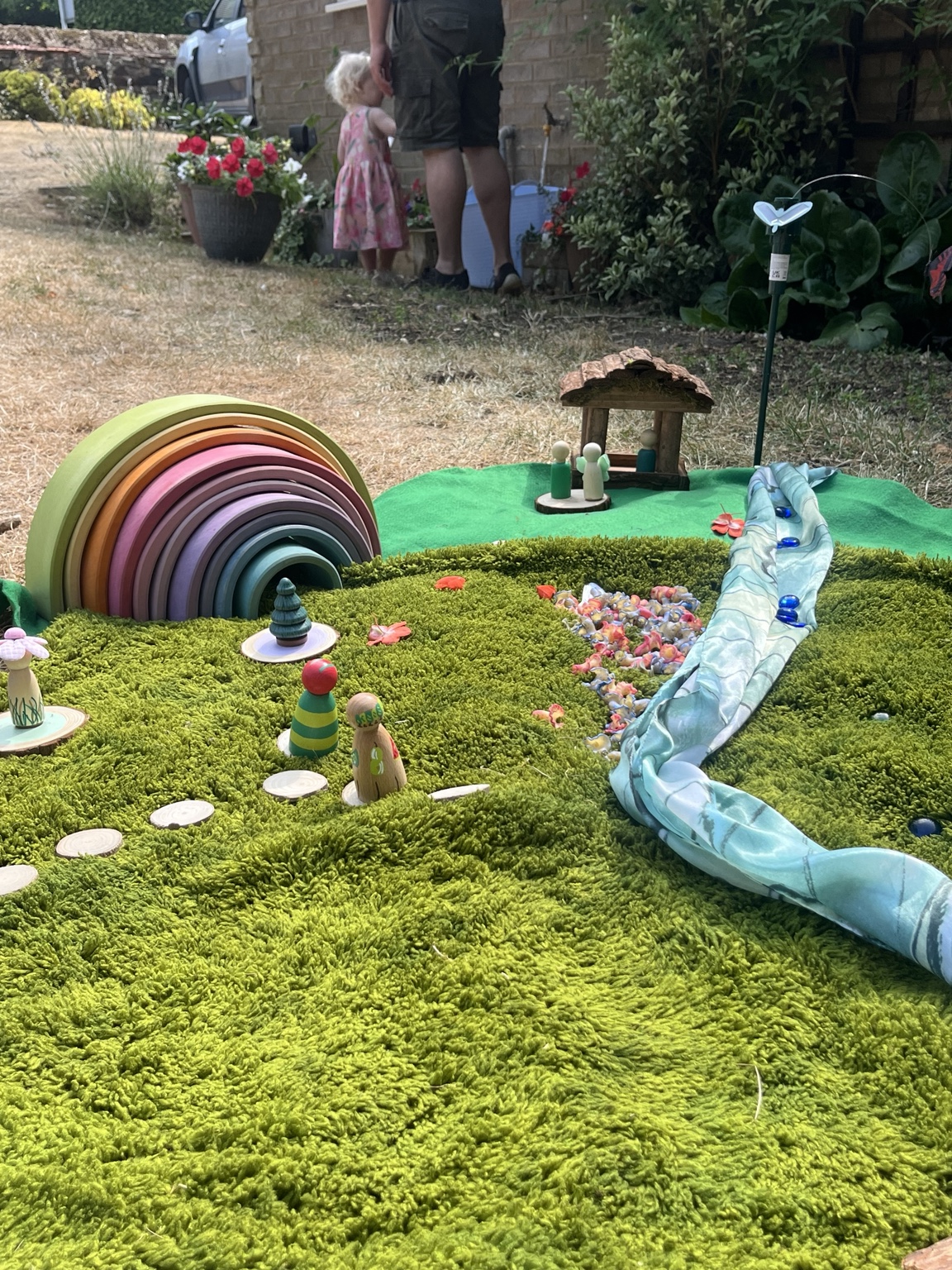 Wooden rainbow, miniature cottage, peg people and a blue fabric stream on a mossy play surface